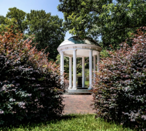 The Old Well. A white, columned gazebo stands at the center of a garden surrounded by lush greenery and manicured bushes. The gazebo has a domed roof and is set on a brick pathway with trees in the background under a clear blue sky. Fearrington Village